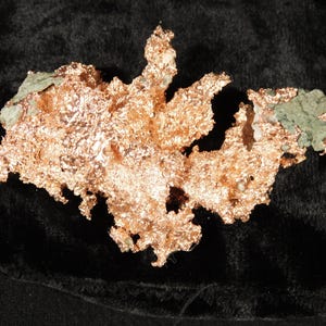 Peut inclure: Un spécimen de cuivre métallique brut présentant une structure cristalline dentelée avec une surface brillante et réfléchissante orange rougeâtre. La formation minérale inclut de petites zones de matrice vert clair terne.