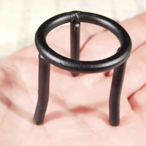 May include: A black metal stand with a circular top and three legs. The stand is designed to hold a small object, such as a crystal or a figurine.