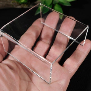 May include: A clear acrylic display stand for small objects, such as crystals or figurines. The stand is square shaped and has a raised platform.