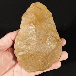 May include: A tan-colored, teardrop-shaped stone tool with a pointed top and a wide, rounded base. The surface features visible flaking and knapped edges, creating a textured, uneven appearance characteristic of a hand-held prehistoric stone artifact.