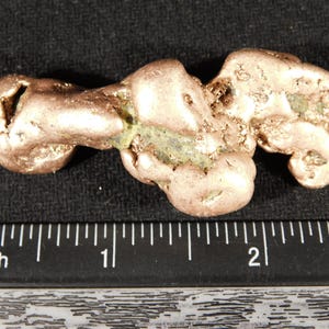 May include: A raw copper nugget with a metallic, reddish-brown surface and irregular, textured formations. The nugget is positioned above a black ruler, showing a length of approximately 2.25 inches from end to end.