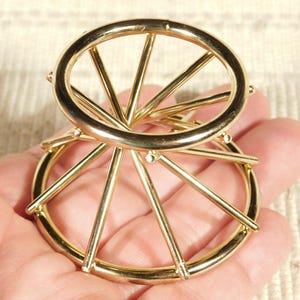 May include: Gold metal ring holder with a circular base and a circular top with spokes. The holder is designed to hold rings upright.