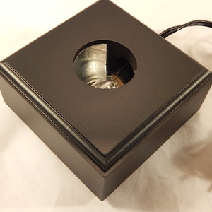 May include: A black, square display base with a circular opening in the center. The base has a beveled edge and a power cord with a plug. The opening reveals a reflective interior, likely for illuminating an object.