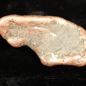 May include: A raw, irregular copper nugget with a metallic, reddish-brown exterior and a textured, grayish-green mineral deposit embedded in the center. The object has a smooth, rounded edge and a rough, uneven surface characteristic of natural ore.