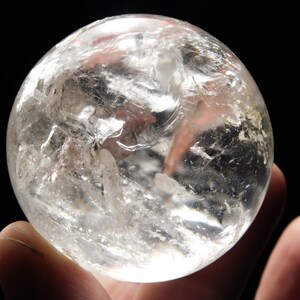 May include: A clear quartz crystal sphere, held in a hand against a black background. The sphere is translucent with internal fractures and inclusions, creating a textured appearance. The light refracts through the crystal, highlighting its clarity.
