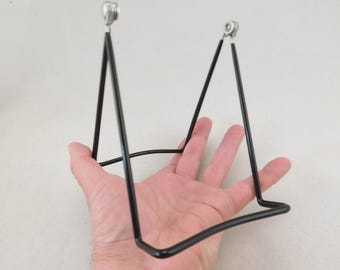 May include: A black metal display stand featuring a triangular frame with two upright supports, a horizontal base, and small silver-toned caps at the top of each vertical arm, designed to hold items in an upright position.
