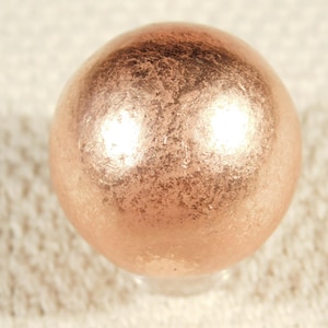May include: A spherical, metallic-looking object with a textured surface and a warm, copper-like color. The sphere appears to be resting on a clear base, and the background is a light, textured fabric.