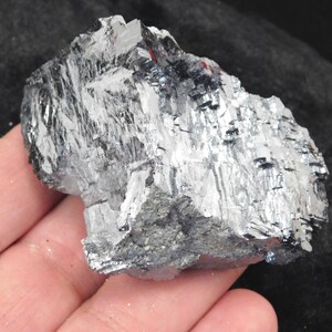 May include: A raw galena mineral specimen featuring a metallic, silver-gray luster with distinct cubic crystalline structures and sharp, angular facets. The mineral displays a heavy, dense appearance with reflective surfaces and a natural, unpolished texture.