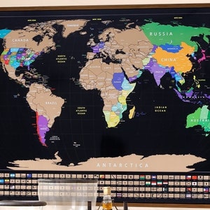 May include: A black scratch-off world map with gold foil accents. The map shows the continents and oceans. The map is framed in a brown wood frame. The text "WORLD TREKKER MAPS" is at the bottom left corner of the map.