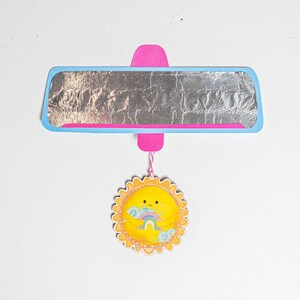 May include: A blue and pink car rearview mirror with a silver reflective surface. A yellow sun with a rainbow and clouds is hanging from the mirror.