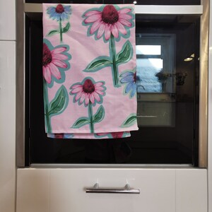 May include: A pink kitchen towel with a floral pattern featuring pink and purple flowers with green leaves. The towel is hanging on a silver oven door.