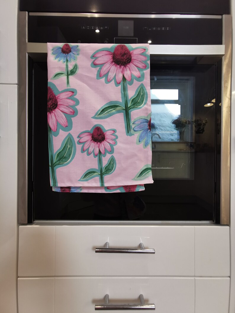 May include: A pink kitchen towel with a floral pattern featuring pink and purple flowers with green leaves. The towel is hanging on a silver oven door.