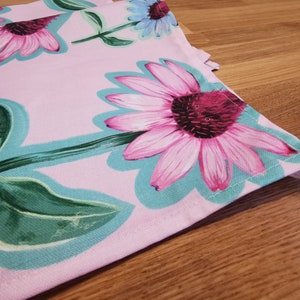 May include: Pink fabric with a floral print featuring pink and green flowers. The fabric has a light blue border and is sewn together with white thread.