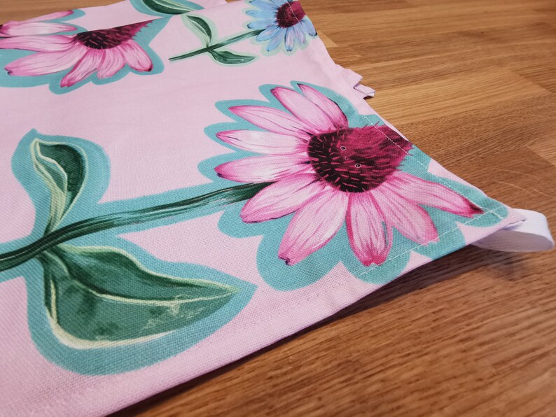 May include: Pink fabric with a floral print featuring pink and green flowers. The fabric has a light blue border and is sewn together with white thread.