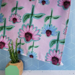 May include: A pink tea towel with a floral pattern of pink and blue flowers. The towel is folded and draped over a blue tiled surface. A green geometric planter with a cactus is in the foreground.