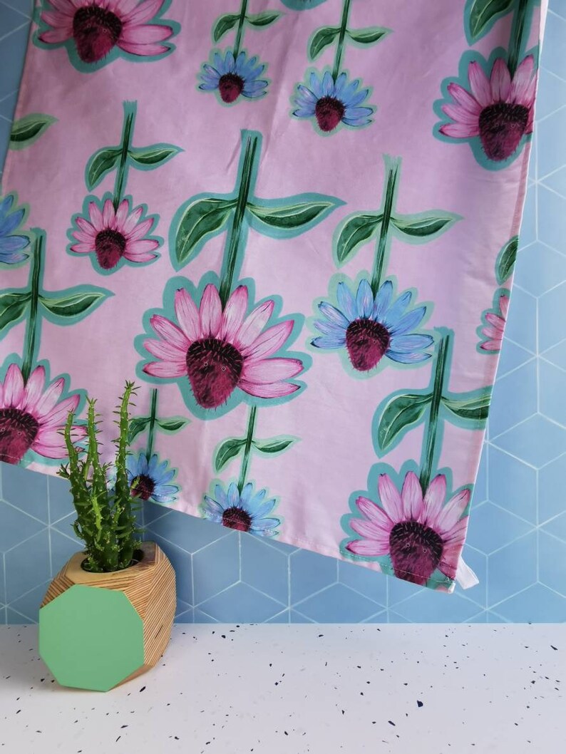 May include: A pink tea towel with a floral pattern of pink and blue flowers. The towel is folded and draped over a blue tiled surface. A green geometric planter with a cactus is in the foreground.