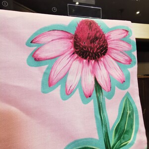 May include: A pink and green kitchen towel with a large pink flower with a red center. The flower has a happy face drawn on it.