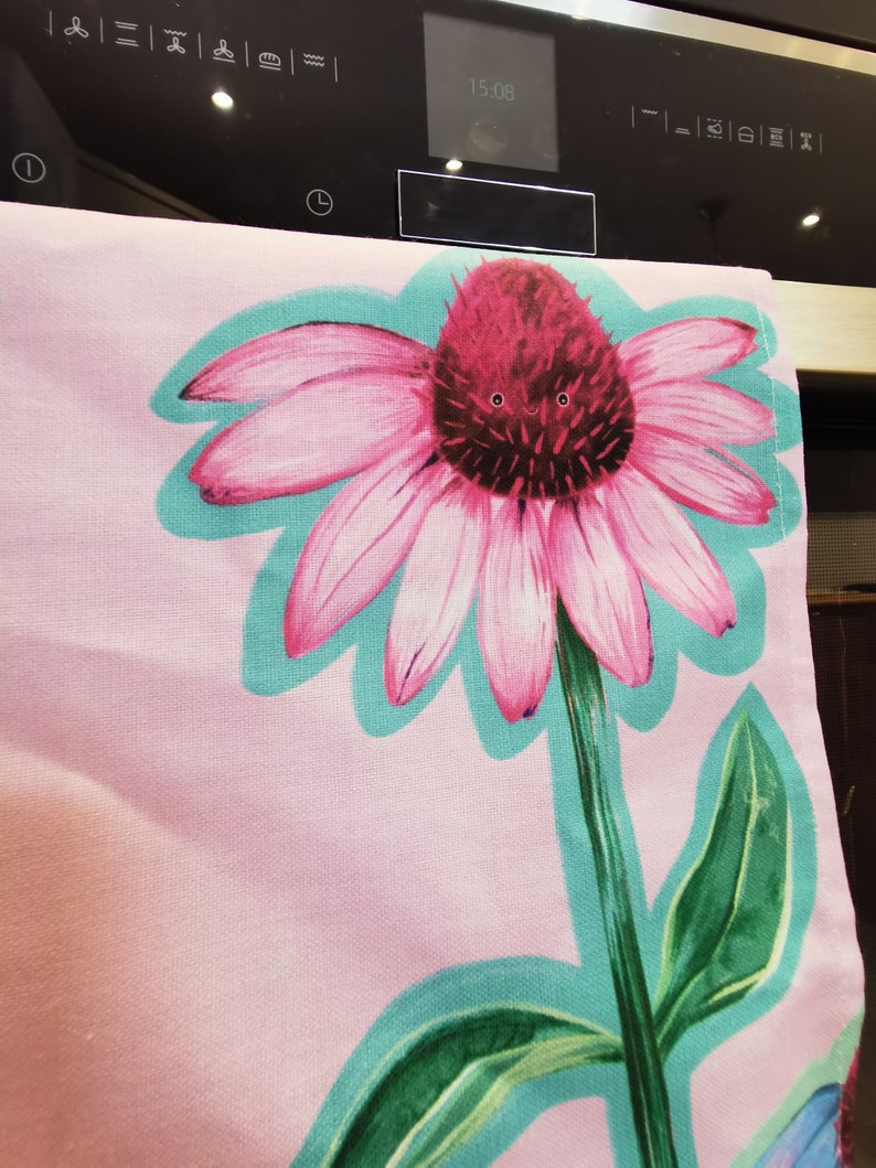 May include: A pink and green kitchen towel with a large pink flower with a red center. The flower has a happy face drawn on it.