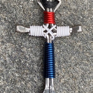 May include: A silver horseshoe cross necklace with red, white, and blue wire wrapped around it. The necklace is made of two horseshoes that are joined together at the top and bottom. The wire is wrapped around the horseshoes in a pattern that resembles the American flag.