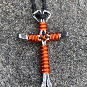 May include: A cross pendant crafted from silver-toned metal horseshoe nails wrapped in bright orange wire. The cross is suspended from a black cord. The design features four nails arranged in a cross shape with wire coils securing the center and arms.