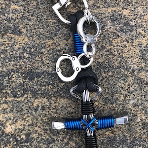 May include: A black leather keychain with a silver handcuff charm and a horseshoe cross made of metal wire wrapped in black and blue thread.