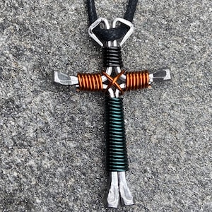 May include: A silver metal cross necklace with a black cord. The cross is made of two horseshoe nails wrapped with copper and green wire.