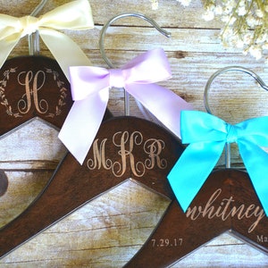 May include: Three wooden hangers with personalized engravings and colorful satin bows. One hanger has the letter "H" inside a wreath, another has the initials "MKR", and the third says "whitney" with "Maid of Honor" below. The bows are cream, lavender, and turquoise.