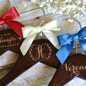 May include: Wooden hangers with personalized text and satin bows in red, cream, and blue. One hanger reads "Jennifer Bridesmaid" with a date. Another features the letter "H" in a wreath, and the third says "Veronica".