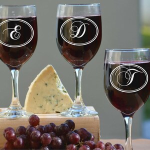 May include: Four wine glasses filled with red wine, each with a different initial inside an oval. A wedge of blue cheese, and a cluster of red grapes sit on a wooden surface. The background is blurred.