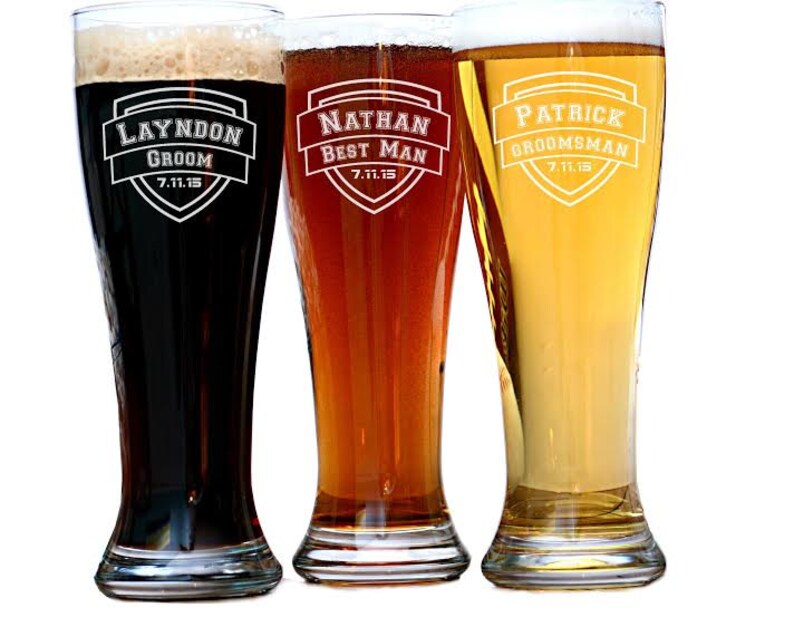 May include: Three tall beer glasses filled with different colored beers. Each glass has a white shield design with a name and title, such as "Layndon Groom" and "Nathan Best Man", along with a date. The beers range from dark to light.