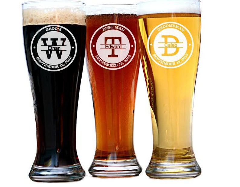 May include: Three personalized pilsner glasses filled with beer. Each glass has a different color of beer and a circular design with text. The first glass says "Groom" and "Ethan", the second says "Best Man" and "Edward", and the third says "Groomsman" and "Caleb".