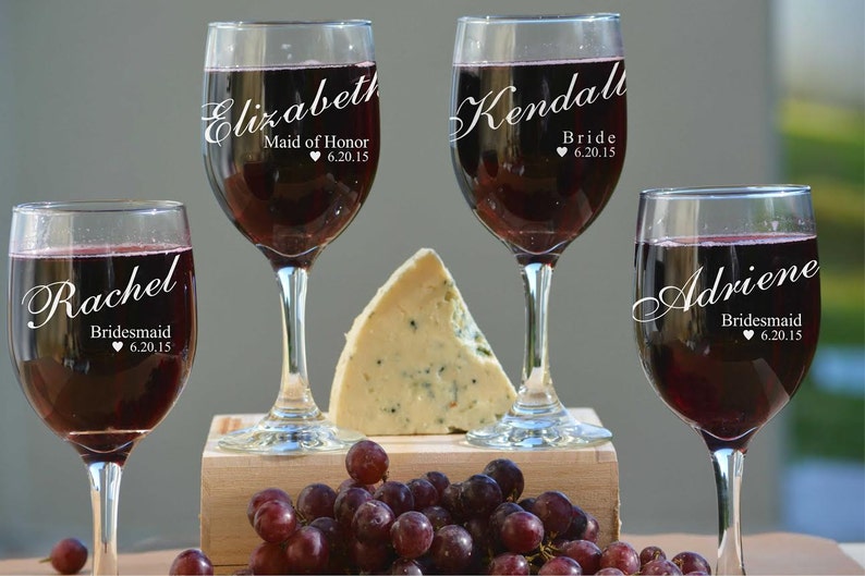 May include: Four wine glasses filled with red wine, each with a name and role printed in white script: Elizabeth, Maid of Honour; Kendall, Bride; Adriene, Bridesmaid; and Rachel, Bridesmaid. A wedge of cheese and grapes are also present.