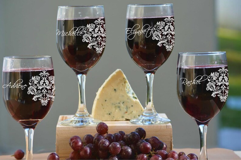 May include: Four wine glasses filled with red wine, each with a different name in elegant script and a white floral design. A wedge of cheese and a cluster of grapes are also present, suggesting a wine and cheese pairing.