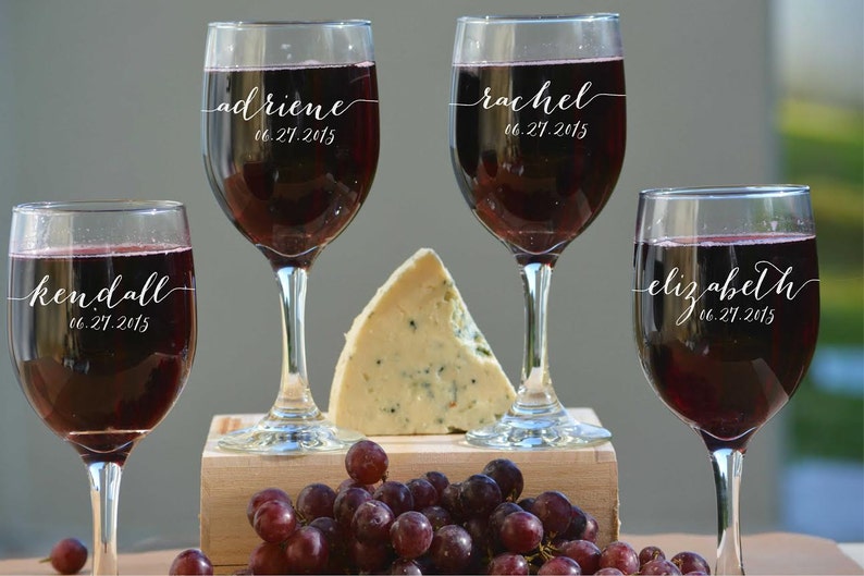 May include: Four wine glasses filled with red wine, each with a name and date in white script: Adriene, Rachel, Kendall, and Elizabeth, with the date 06.27.2015. A wedge of cheese and grapes are on a wooden surface.