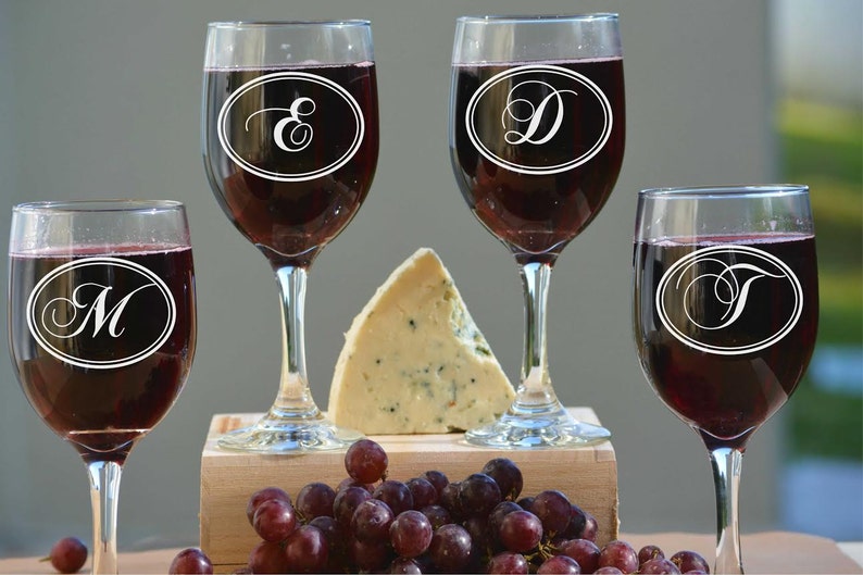 May include: Four wine glasses filled with red wine, each with a different initial inside an oval. A wedge of blue cheese, and a cluster of red grapes sit on a wooden surface. The background is blurred.