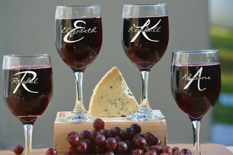 May include: Four wine glasses filled with red wine, each with a different name etched on the glass: Elizabeth, Kendall, Rachel, and Adriene. A wedge of blue cheese and a cluster of red grapes are in the center.