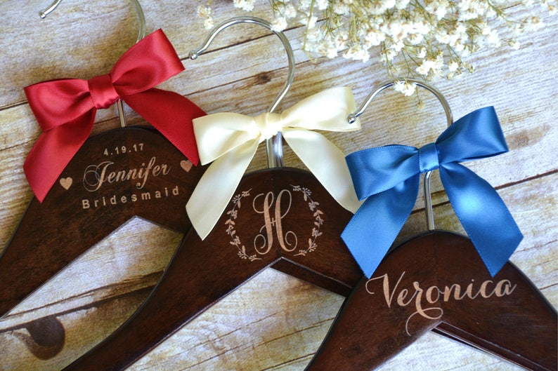 May include: Wooden hangers with personalized text and satin bows in red, cream, and blue. One hanger reads "Jennifer Bridesmaid" with a date. Another features the letter "H" in a wreath, and the third says "Veronica".