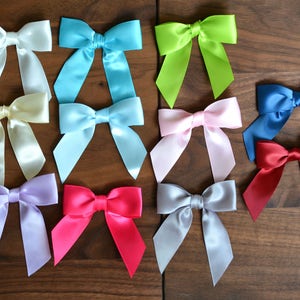 May include: A collection of satin bows in various colors, including white, light blue, green, pink, blue, and red. The bows are arranged on a wooden surface, showcasing their vibrant hues and elegant design. Perfect for crafting or gift wrapping.