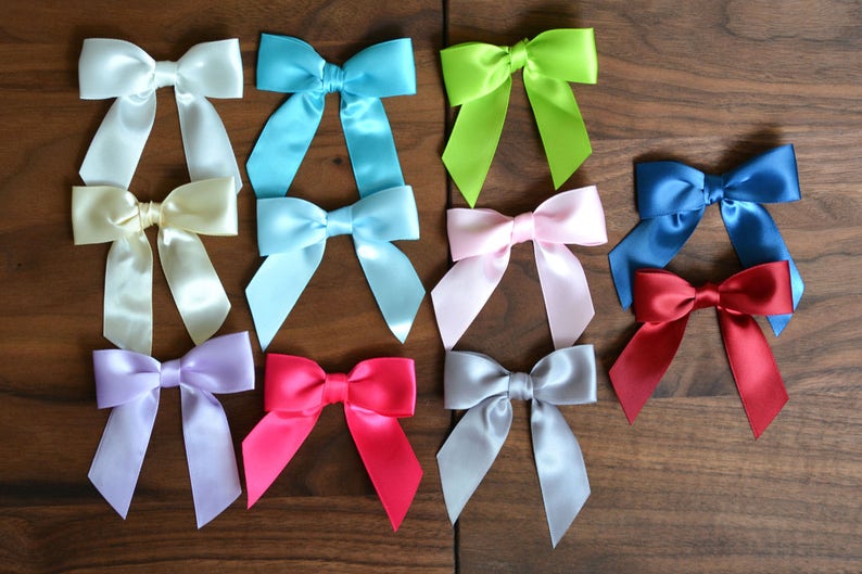 May include: A collection of satin bows in various colors, including white, light blue, green, pink, blue, and red. The bows are arranged on a wooden surface, showcasing their vibrant hues and elegant design. Perfect for crafting or gift wrapping.