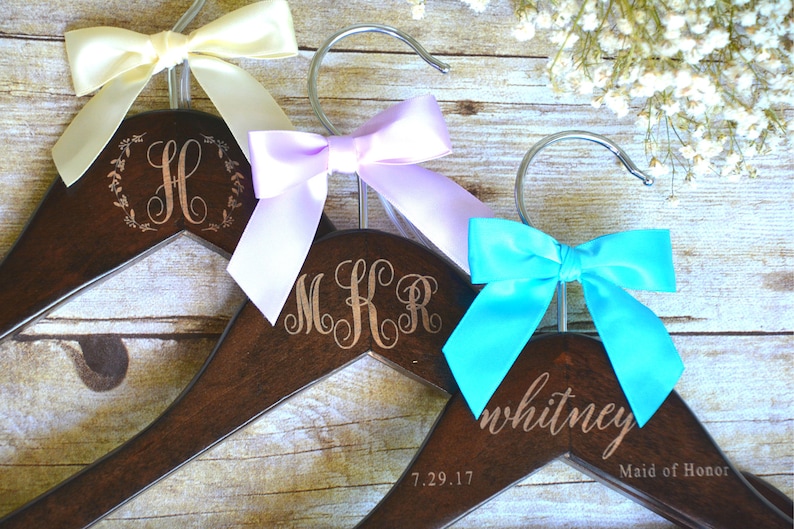 May include: Three wooden hangers with personalized engravings and colorful satin bows. One hanger has the letter "H" inside a wreath, another has the initials "MKR", and the third says "whitney" with "Maid of Honor" below. The bows are cream, lavender, and turquoise.