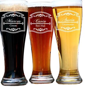 May include: Three tall beer glasses filled with different colored beers: dark, amber, and golden. Each glass has a decorative white label with a name and title: Marcus, Groom; Gavin, Best Man; and Austin, Groomsman. The glasses are on a white background.