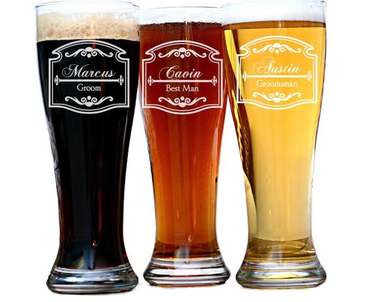 May include: Three tall beer glasses filled with different colored beers: dark, amber, and golden. Each glass has a decorative white label with a name and title: Marcus, Groom; Gavin, Best Man; and Austin, Groomsman. The glasses are on a white background.