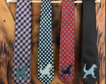 May include: Four neckties hanging on a wooden hanger. The ties are all different patterns and colors, and each has a dinosaur applique on the bottom. The ties are a plaid pattern in red and blue, a black and white pattern with circles, a red and black pattern with small flowers, and a black and white polka dot pattern.
