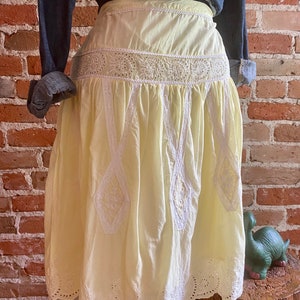 May include: A yellow cotton skirt with white embroidery and lace trim. The skirt has a high waist and a full, flowing silhouette.