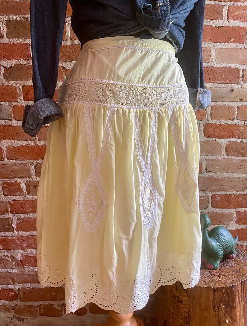 May include: A yellow cotton skirt with white embroidery and lace trim. The skirt has a high waist and a full, flowing silhouette.