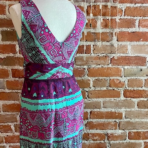 Peut inclure: Une robe sans manches avec un d&eacute;collet&eacute; en V et un motif g&eacute;om&eacute;trique dans des tons de rose, violet et turquoise. La robe est sur un mannequin devant un mur de briques.