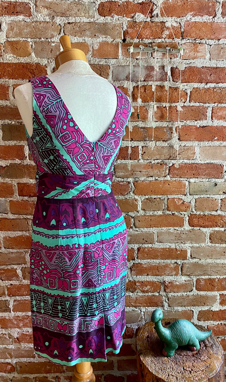 Peut inclure: Une robe sans manches avec un d&eacute;collet&eacute; en V et un motif g&eacute;om&eacute;trique dans des tons de rose, violet et turquoise. La robe est sur un mannequin devant un mur de briques.