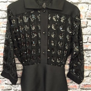 Op de afbeelding: Een zwart paillettenblouse met een knoopsluiting aan de voorkant en een getailleerde pasvorm. Het blouse heeft een transparante, paillettenoverleg en een effen zwarte onderkant.
