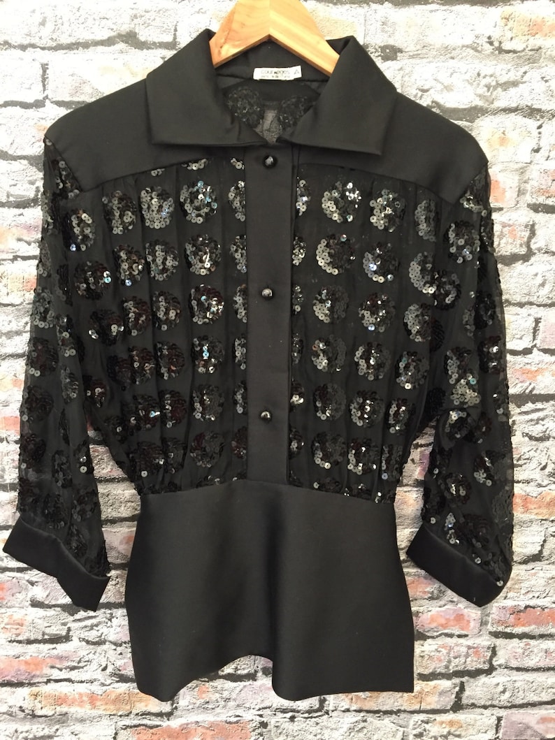 Op de afbeelding: Een zwart paillettenblouse met een knoopsluiting aan de voorkant en een getailleerde pasvorm. Het blouse heeft een transparante, paillettenoverleg en een effen zwarte onderkant.