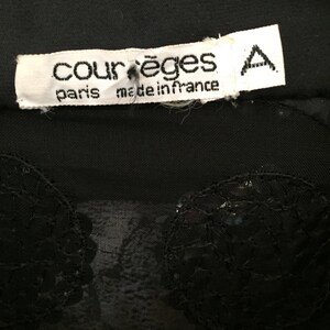 Op de afbeelding: Zwart doorschijnend topje met een zwarte kanten overlay en een wit label met de tekst "Courr&egrave;ges Paris made in France" met een hoofdletter "A" gedrukt aan de rechterkant van het label.